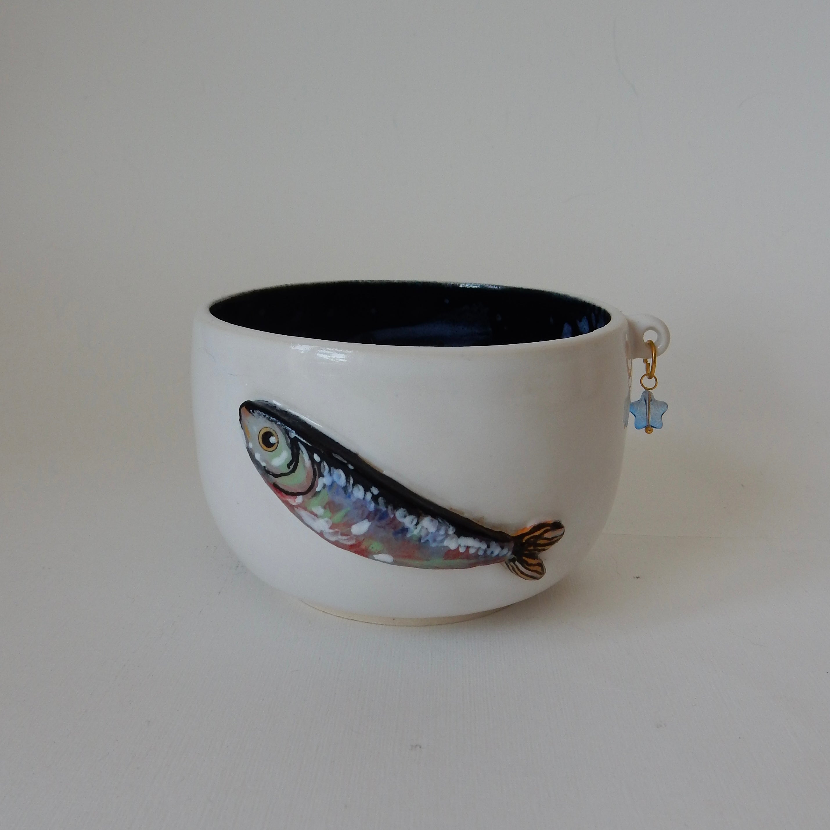 A white ceramic bowl with a dark blue interior, featuring a hand-painted fish design on the front and a small blue hanging charm, set against a plain light background.