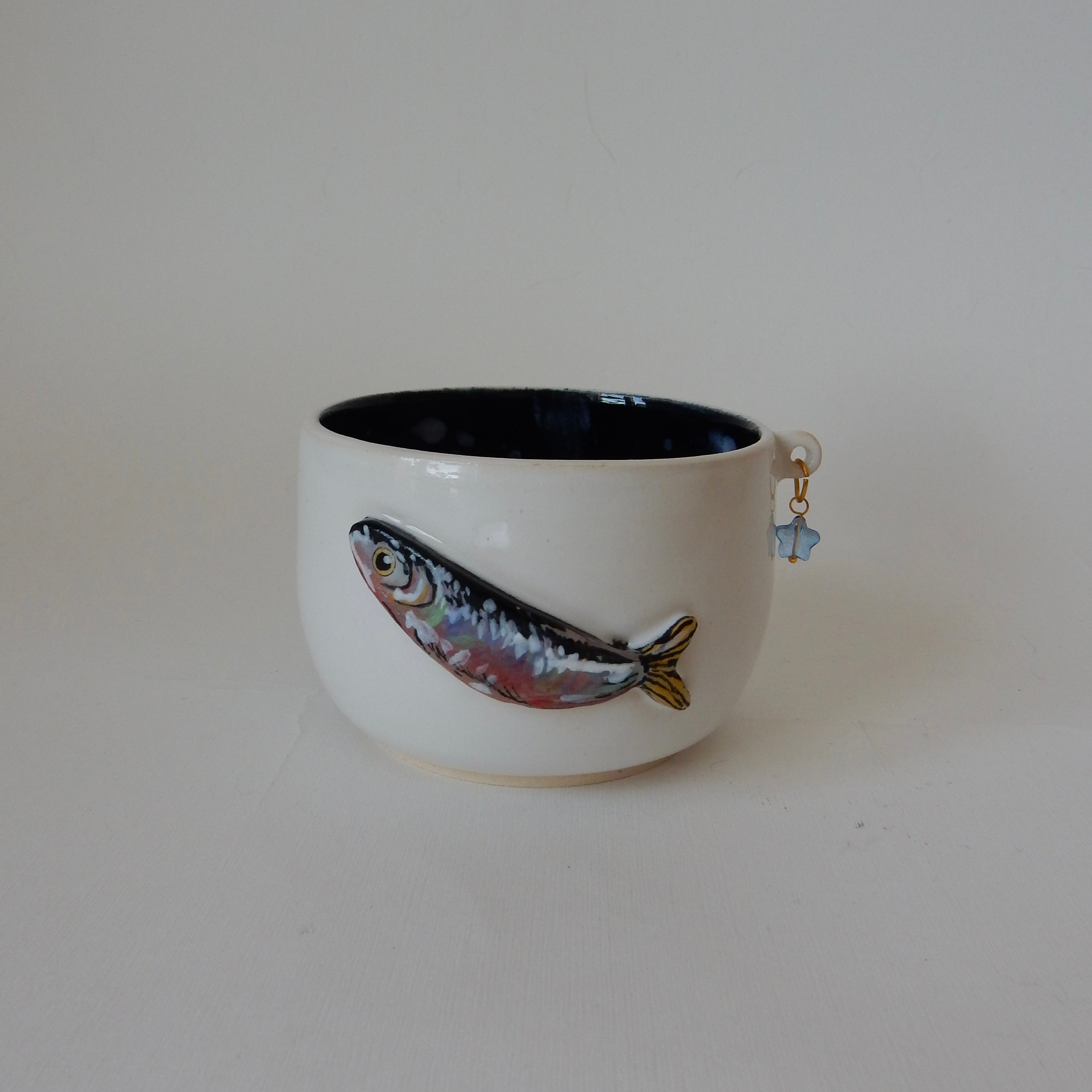 A white ceramic bowl with a dark blue interior, featuring a hand-painted fish design on the front and a small blue hanging charm, set against a plain light background.